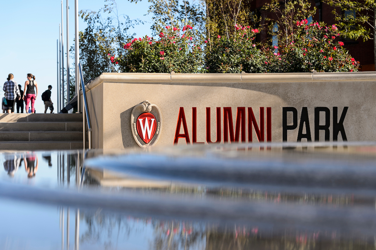 W crest and text, Alumni Park, adorn wall covered with flowers and fountain in foreground.