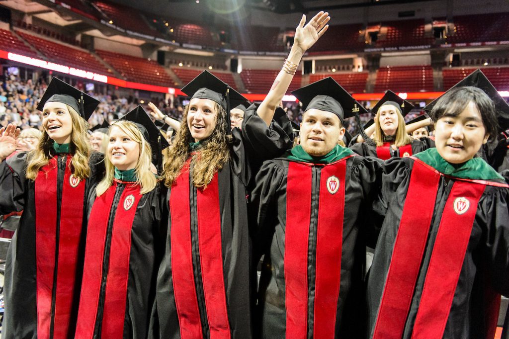 Group of graduates in academic attire standing side-by-side smiling while singing Varsity.