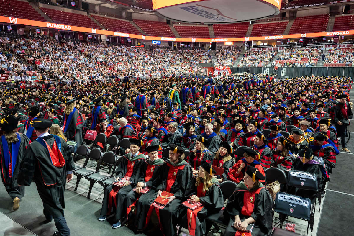Graduates in academic attire seated in rows at the Kohl Center, surrounded by families and guests.